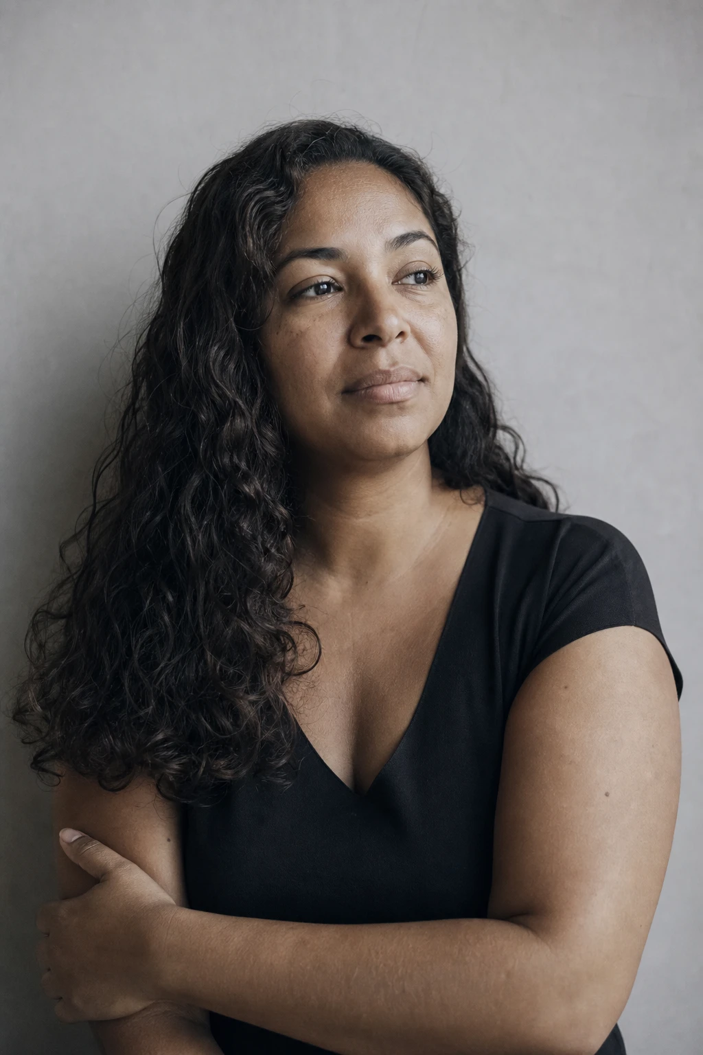 Retrato editorial de uma mulher de braços cruzados diante de uma parede neutra, expressão de força tranquila e serena, tratamento sóbrio em tons frios